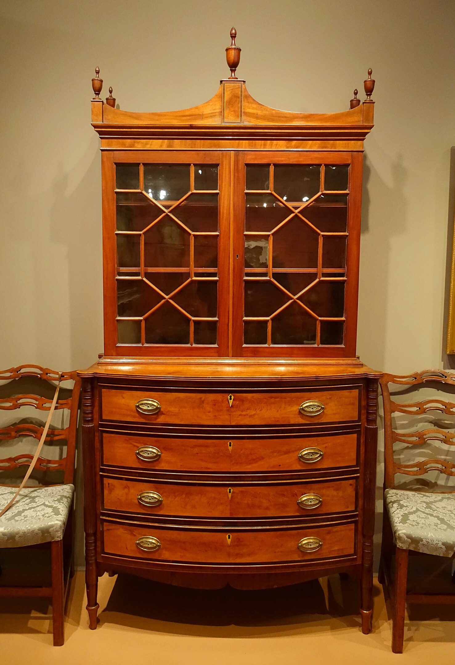 Antique wooden secretary bookcase with bow-front drawers, glass cabinet doors featuring geometric mullions, decorative finials atop the crown, flanked by two matching ladder-back chairs with patterned upholstered seats in a museum setting