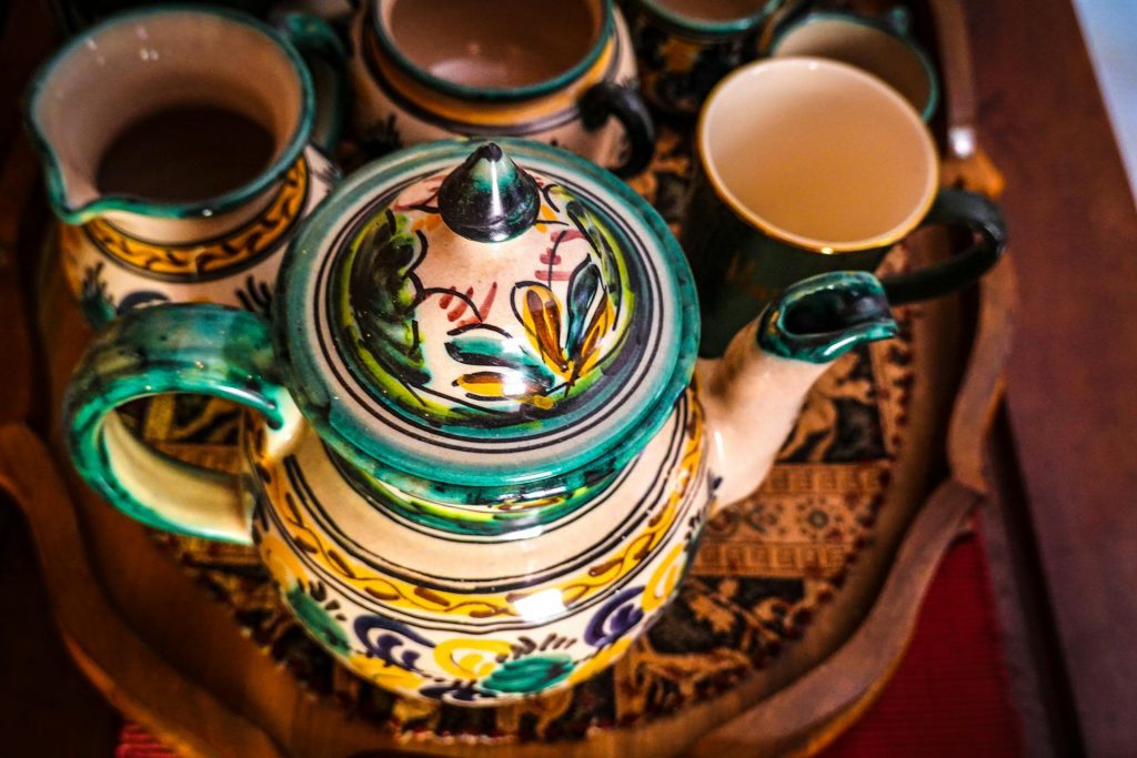 Colorful ceramic teapot set with matching cups, decorated with traditional patterns and vivid green, yellow, and brown tones, arranged on a round wooden tray with carvings, viewed from above, likely part of a cultural or decorative tea service