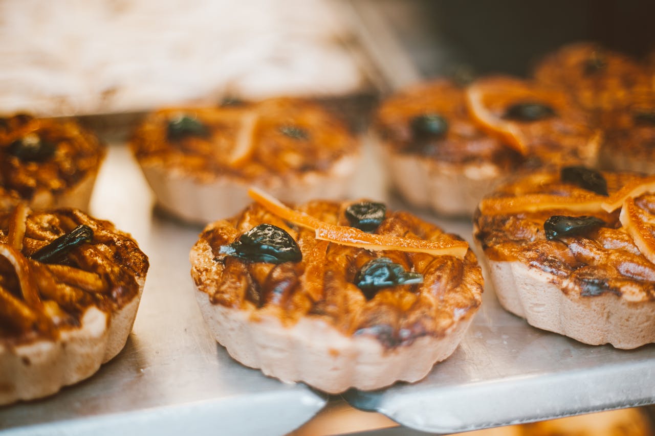 Close-up of mini fruit pies with golden crust, orange peels, dark fruit toppings, arranged on a metal tray