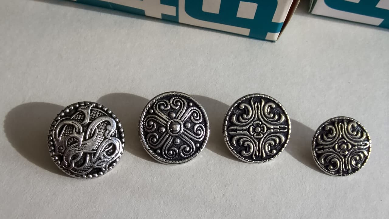 Alt description (34 words): Four ornate silver-tone metal buttons arranged in a row on white paper. Each circular button features different intricate Celtic or medieval-inspired designs with swirls, curves, and decorative patterns. A box is partially visible at the top