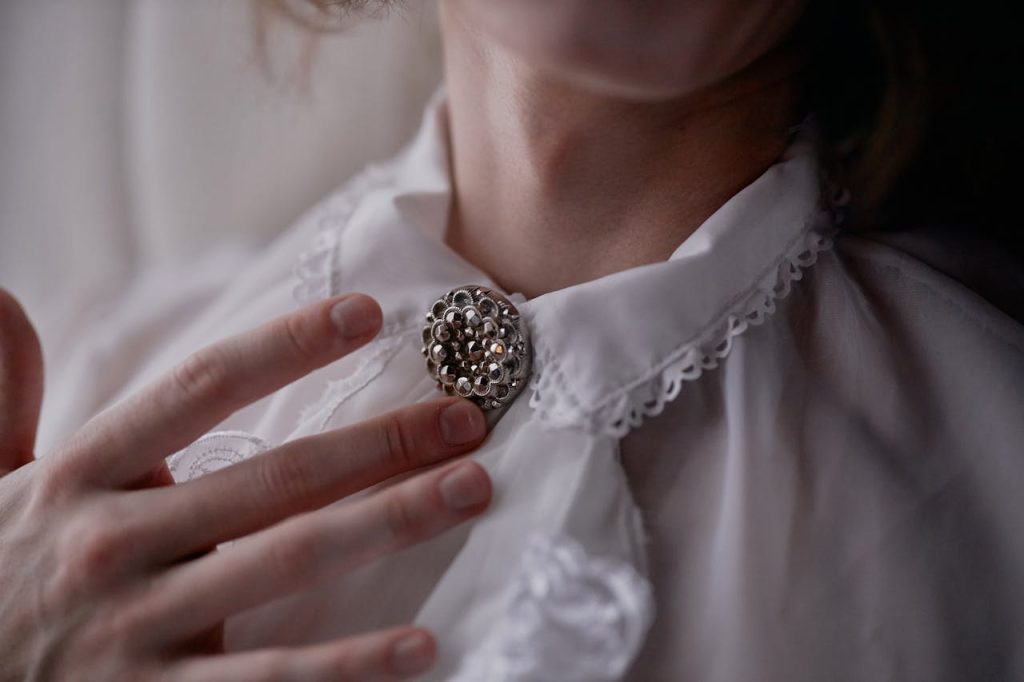 Close-up of a person wearing a white lace-collared blouse, vintage silver brooch pinned at the neck, fingers gently touching brooch