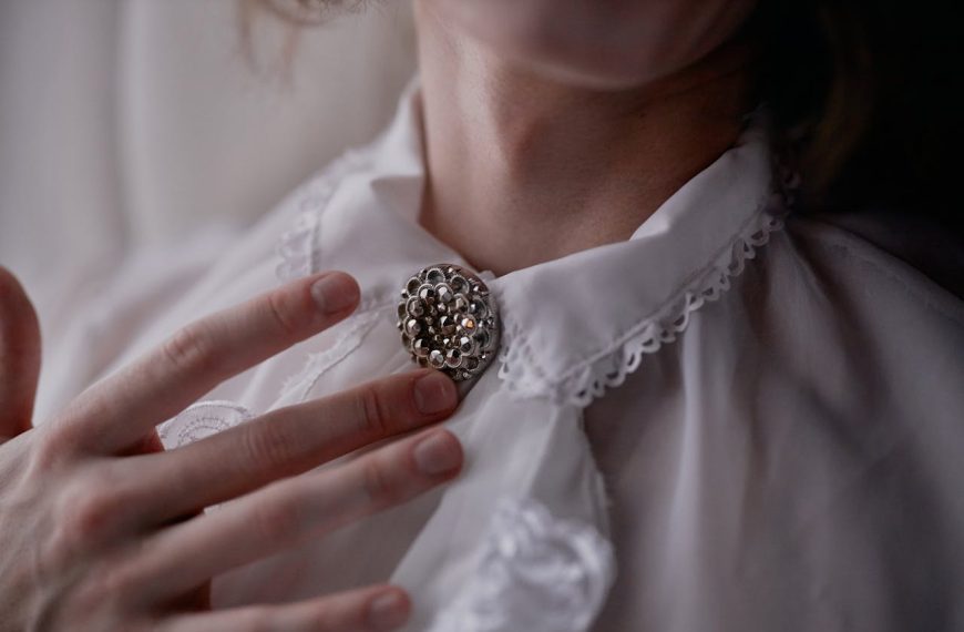 Close-up of a person wearing a white lace-collared blouse, vintage silver brooch pinned at the neck, fingers gently touching brooch