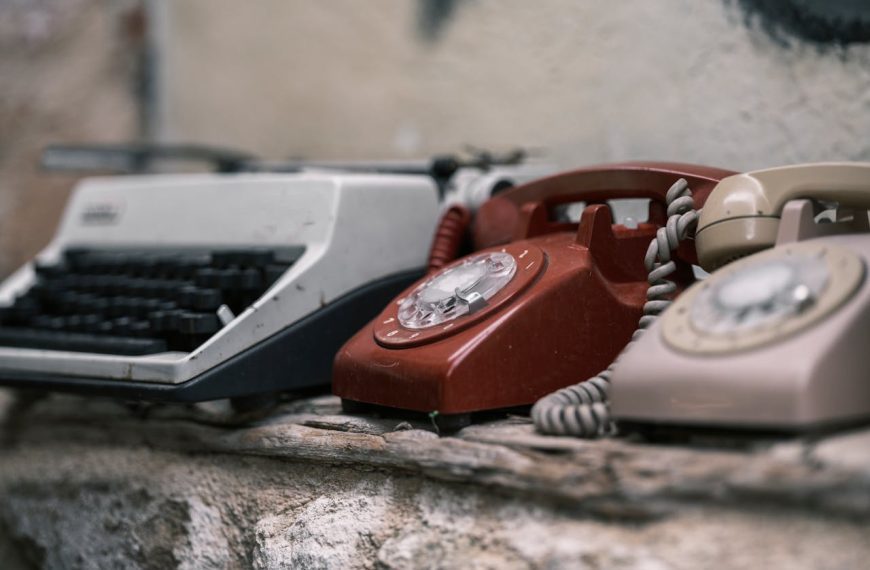 Vintage typewriter, red rotary phone, and beige rotary phone placed on a weathered stone surface against a faded wall