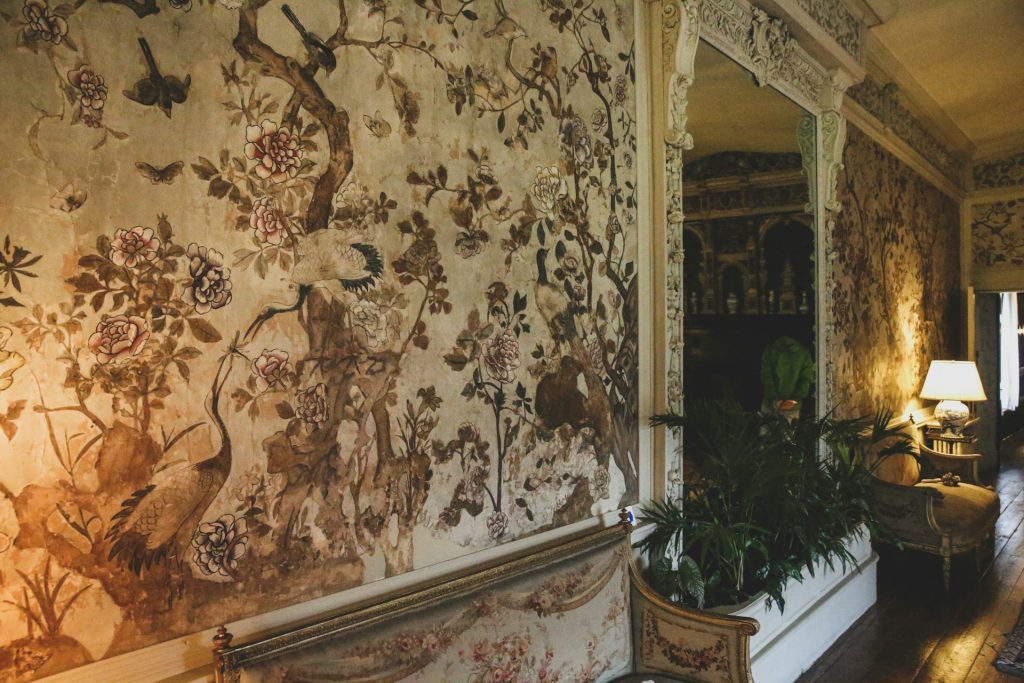 antique hallway, distressed floral wallpaper, vintage interior design, ornate white trim, classic furniture, dim lamp lighting, elegant and aged atmosphere