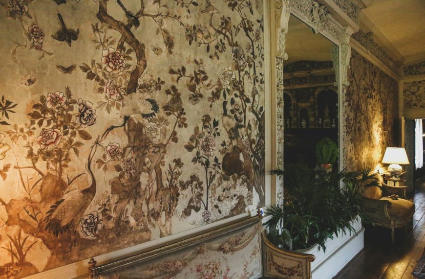 antique hallway, distressed floral wallpaper, vintage interior design, ornate white trim, classic furniture, dim lamp lighting, elegant and aged atmosphere