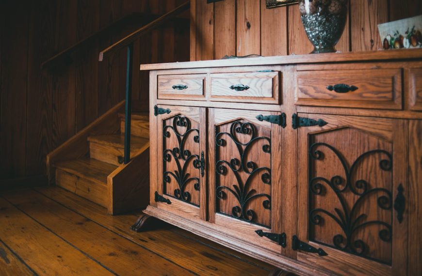 Wooden cabinet with ornate black metal detailing, rustic interior setting, placed beside wooden staircase, warm natural lighting, polished wood floor, vintage-style decor
