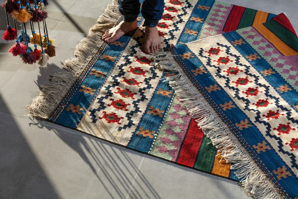 Colorful woven rug, traditional geometric patterns, fringe edges, person standing barefoot, sunlight casting shadows