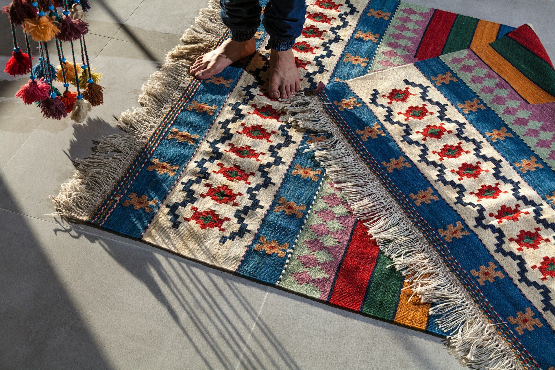 Colorful woven rug, traditional geometric patterns, fringe edges, person standing barefoot, sunlight casting shadows