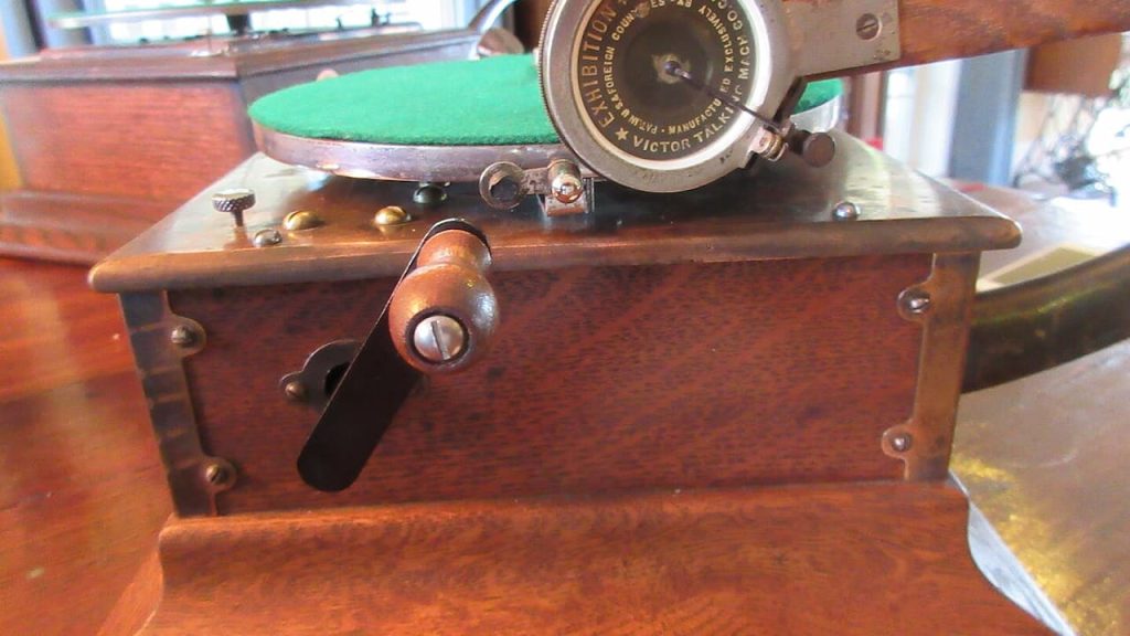 Antique Victor Talking Machine phonograph with wooden cabinet, metal crank handle, and circular metal reproducer. The turntable features a green felt platter surface. The vintage record player sits on a polished wooden table near a window