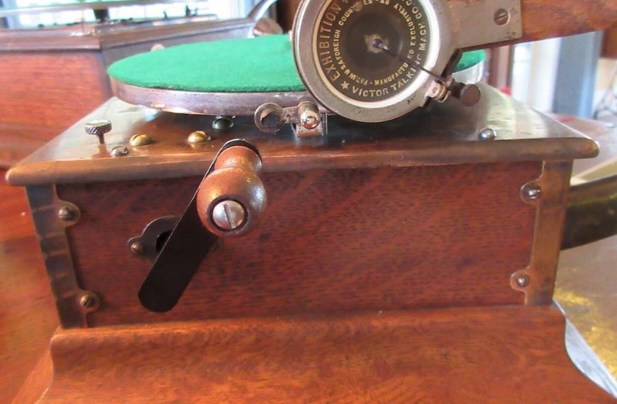 Antique Victor Talking Machine phonograph with wooden cabinet, metal crank handle, and circular metal reproducer. The turntable features a green felt platter surface. The vintage record player sits on a polished wooden table near a window