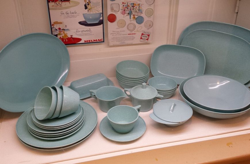 A collection of vintage light blue Melmac dinnerware, including plates, bowls, cups, saucers, serving dishes, and lidded containers, arranged neatly on white shelves with colorful retro advertisements displayed on the wall behind them