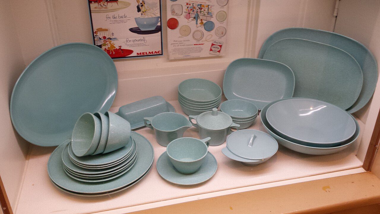 A collection of vintage light blue Melmac dinnerware, including plates, bowls, cups, saucers, serving dishes, and lidded containers, arranged neatly on white shelves with colorful retro advertisements displayed on the wall behind them