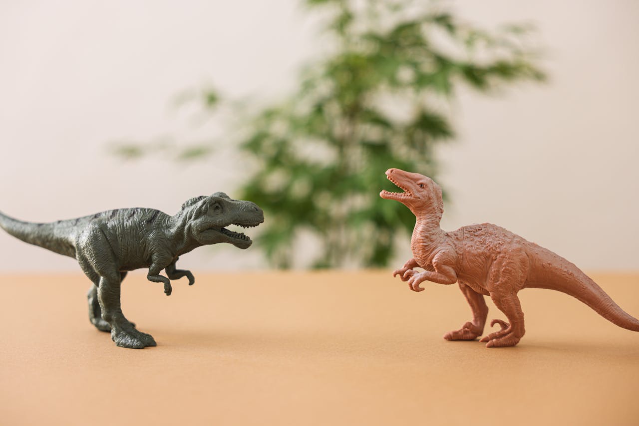 Two plastic dinosaur toys, green Tyrannosaurus rex, brown Velociraptor, posed on a flat surface, blurred plant background