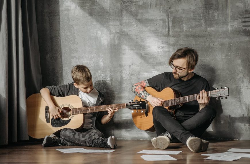 Child and man sitting on floor playing acoustic guitars, sheet music scattered, casual indoor music practice, gray wall background