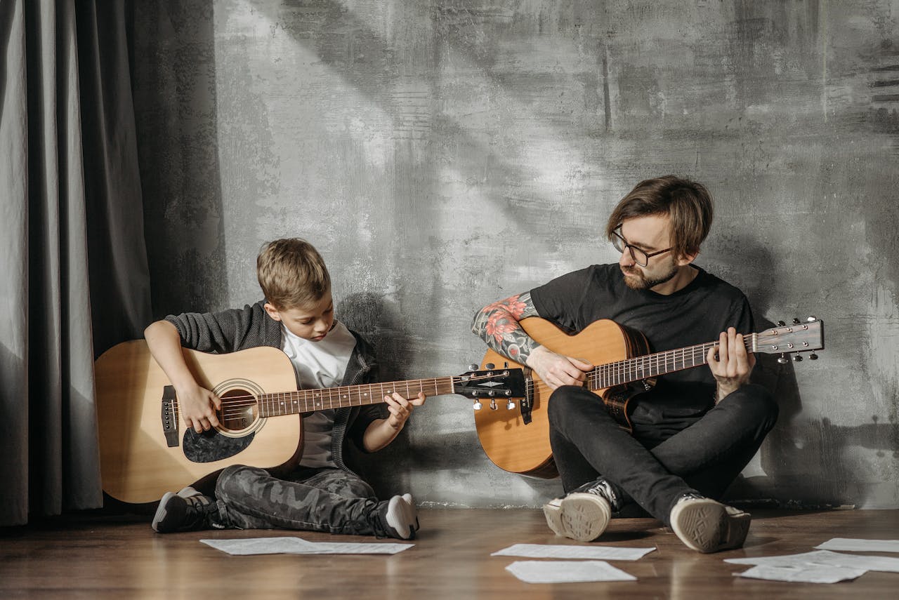 Child and man sitting on floor playing acoustic guitars, sheet music scattered, casual indoor music practice, gray wall background