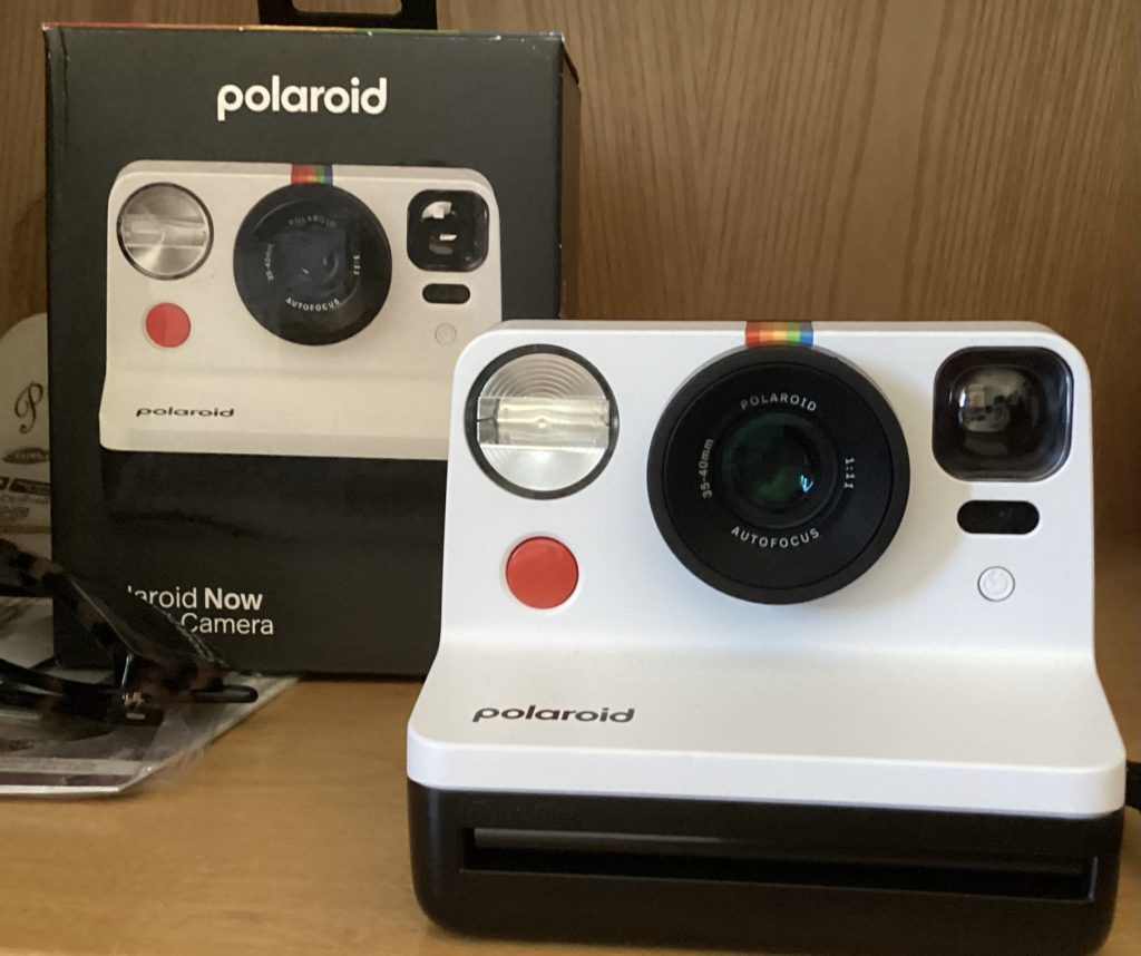 White Polaroid Now instant camera placed in front of its black retail packaging, displayed on a wooden shelf