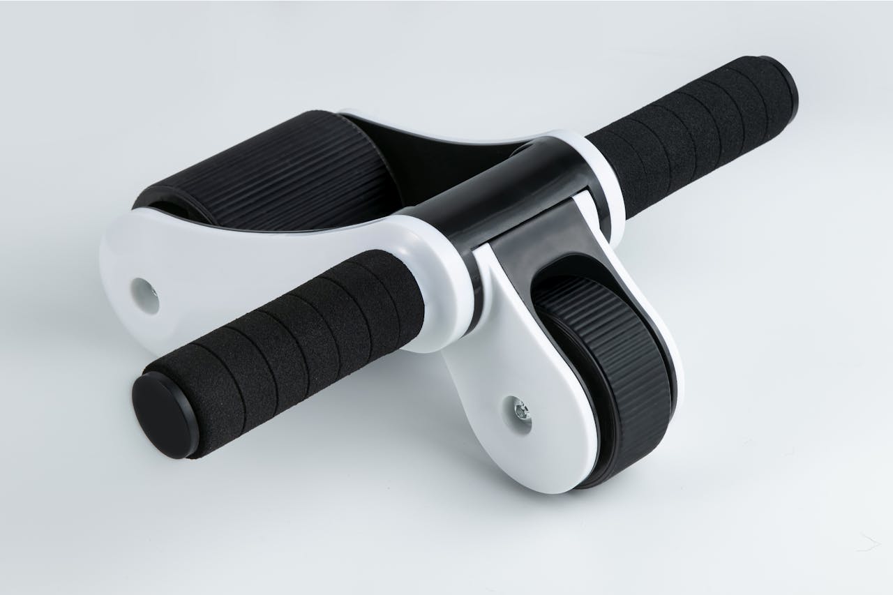 An ab roller exercise equipment with black foam handles and a white plastic frame. The central wheel is black with textured grip. The device is photographed against a plain light gray background in studio lighting