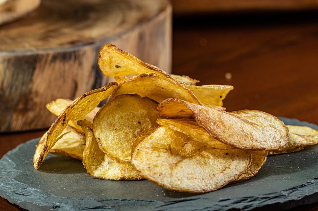 Golden crispy potato chips, slightly curled edges, served on a dark slate plate, rustic wooden background