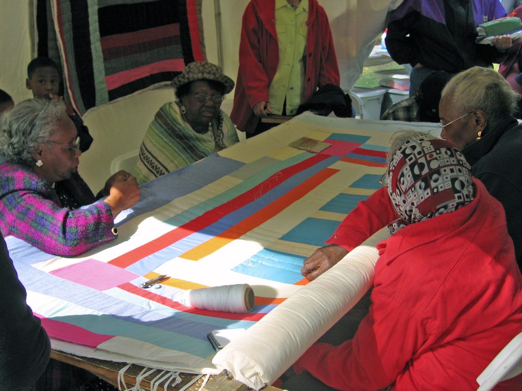 group of elderly women quilting together, colorful patchwork fabric on large table, quilts hanging in background