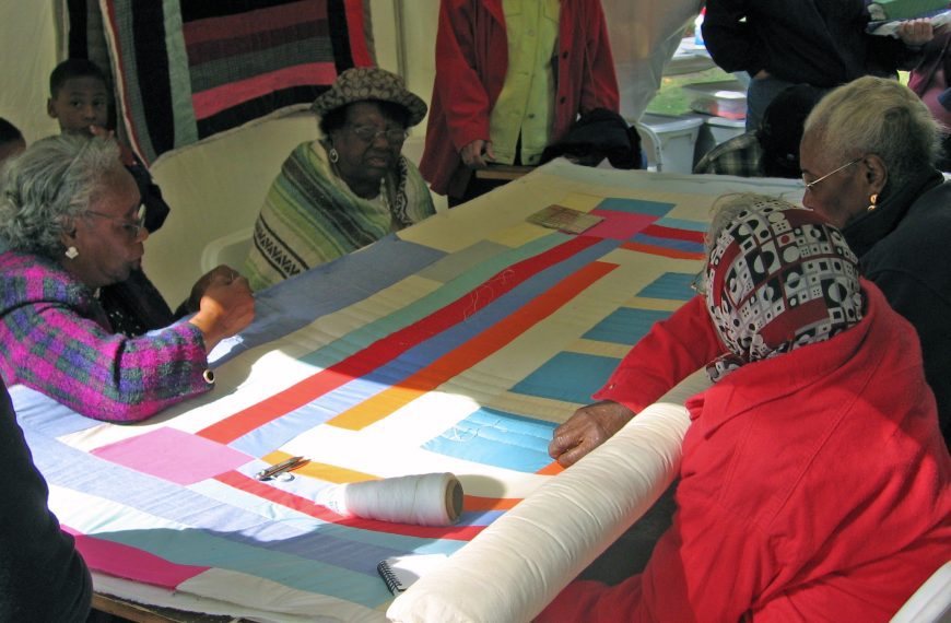 group of elderly women quilting together, colorful patchwork fabric on large table, quilts hanging in background