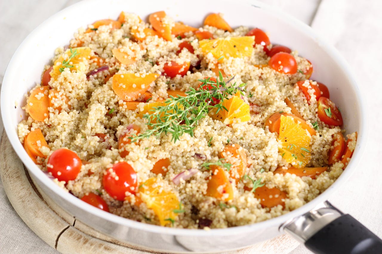 Colorful quinoa salad in a white pan, mixed with cherry tomatoes, orange slices, carrots, fresh herbs, served on a wooden board