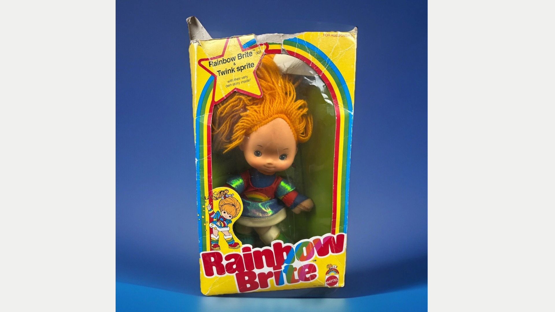 Rainbow Brite doll in original yellow packaging with visible rainbow design and character artwork, doll has orange spiky hair