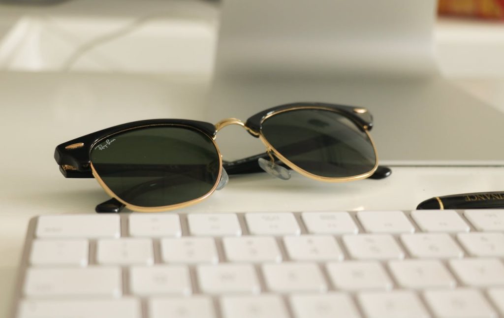 Ray-Ban Wayfarer sunglasses with black and gold frame resting on a white desk near a keyboard and pen
