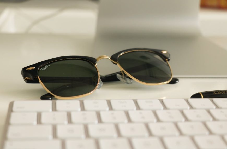 Ray-Ban Wayfarer sunglasses with black and gold frame resting on a white desk near a keyboard and pen