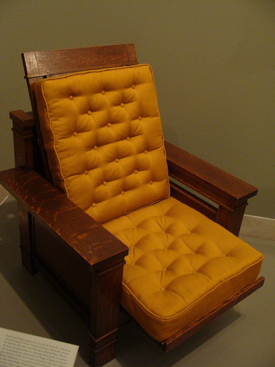 Arts and Crafts style wooden reclining armchair with mustard yellow tufted cushions on both seat and backrest. Features wide wooden armrests, reclining mechanism, and footrest extension, displayed in a museum setting against a beige wall