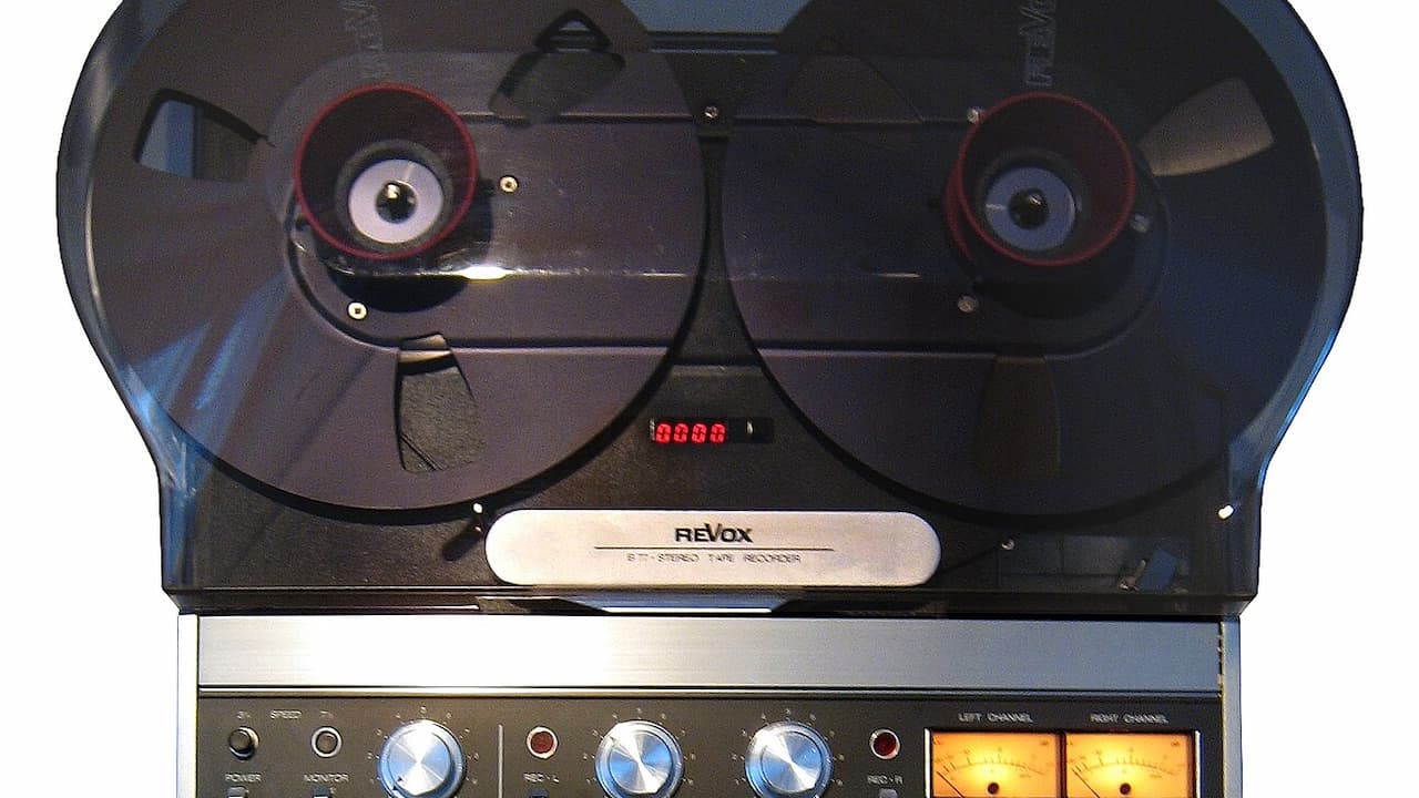 Close-up of a Revox reel-to-reel tape recorder showing two tape reels with red centers, a digital counter displaying "0000", and control panel with multiple knobs and illuminated VU meters at bottom