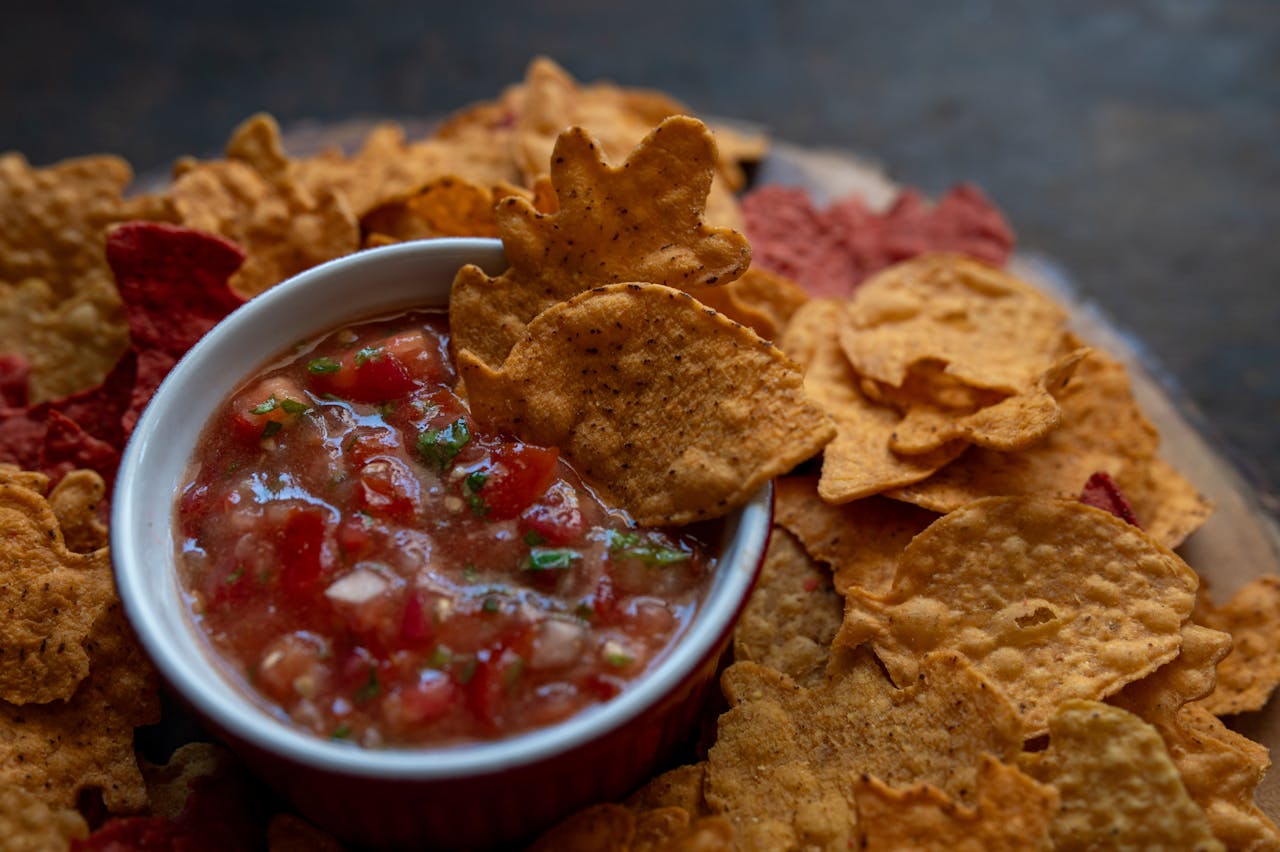Bowl of chunky red salsa, surrounded by leaf-shaped tortilla chips, close-up view, snack setup on dark background