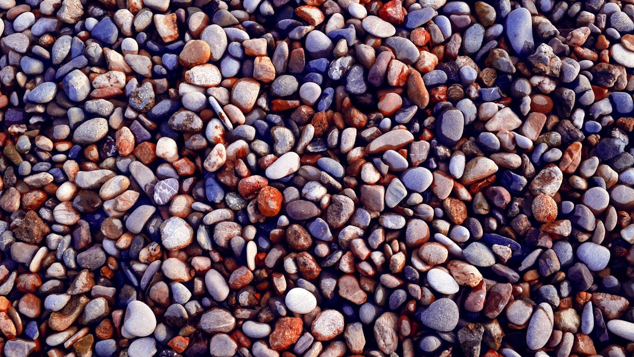 A dense collection of smooth river pebbles and stones in various colors including gray, white, rust, and purple-blue. The stones are polished and rounded from water erosion, creating a textured natural mosaic that fills the entire frame