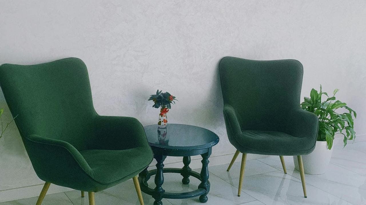 Two green upholstered chairs with wooden legs, small round dark green table between them, small vase with flowers on the table, white textured wall background, white tiled floor, potted green plant to the right