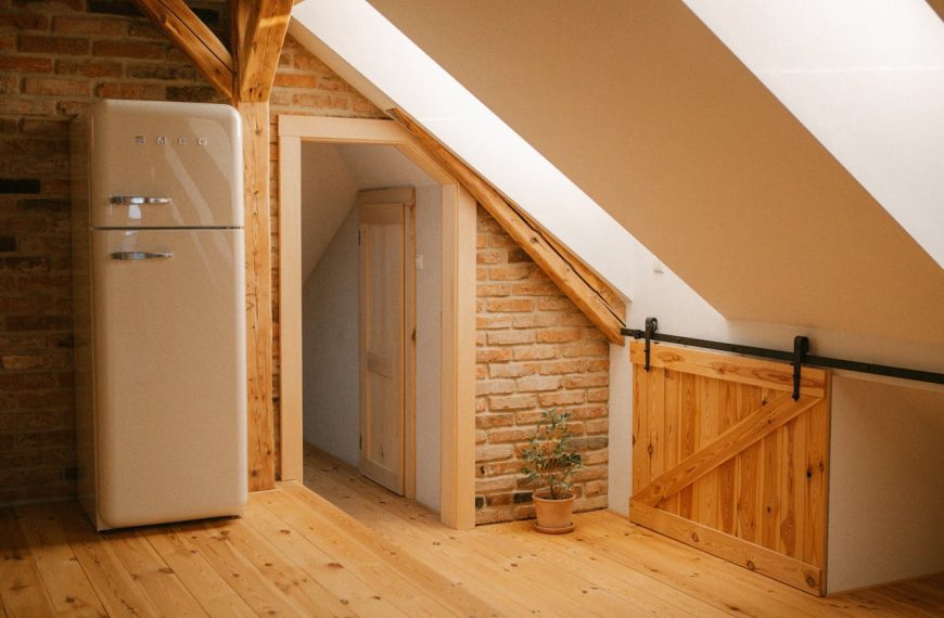 Rustic attic kitchen space with exposed brick wall, wooden pine flooring, and sloped ceiling. Features retro cream SMEG refrigerator, sliding barn door on metal track, small potted plant, and doorway leading to another room