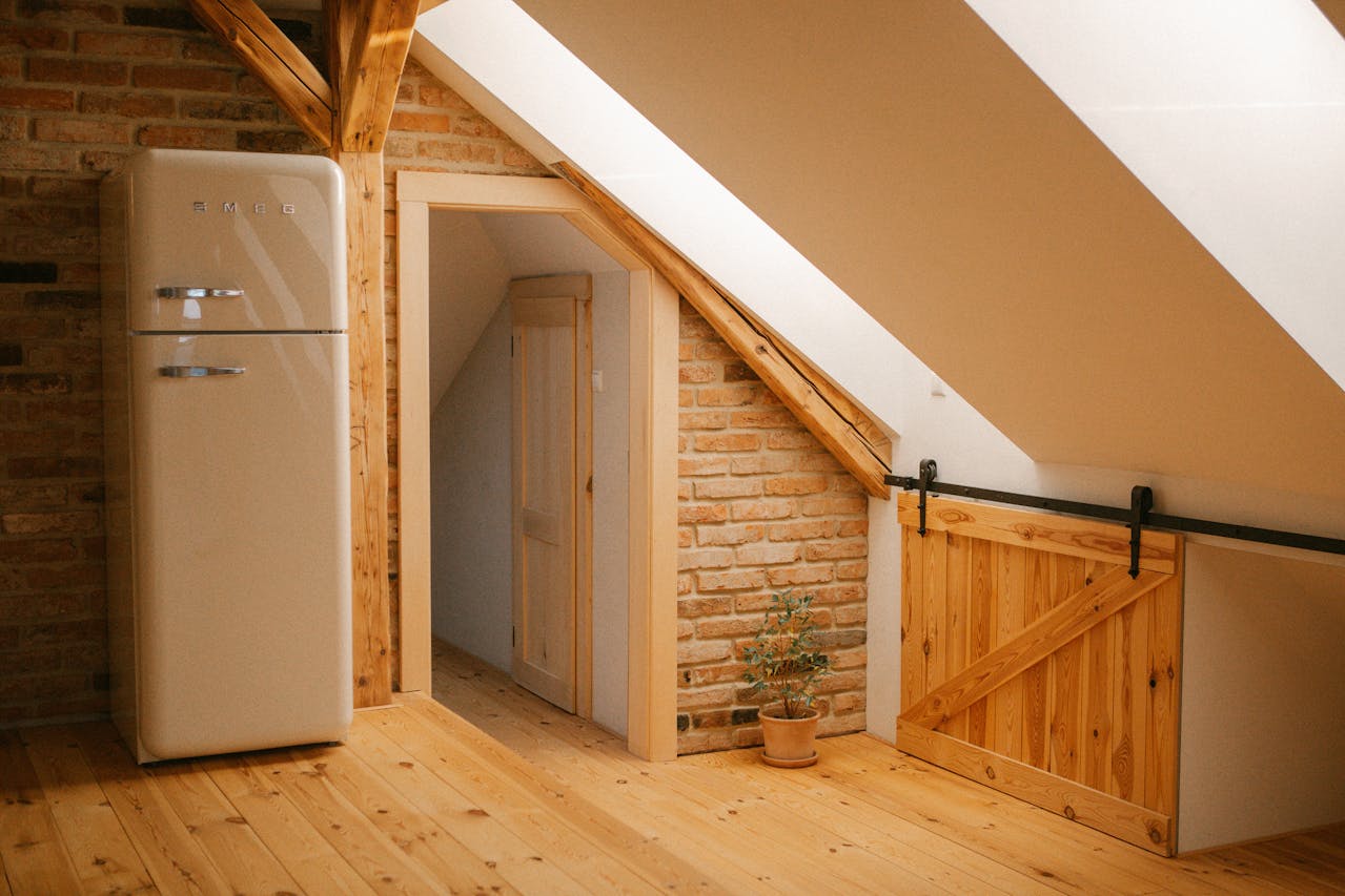 Rustic attic kitchen space with exposed brick wall, wooden pine flooring, and sloped ceiling. Features retro cream SMEG refrigerator, sliding barn door on metal track, small potted plant, and doorway leading to another room