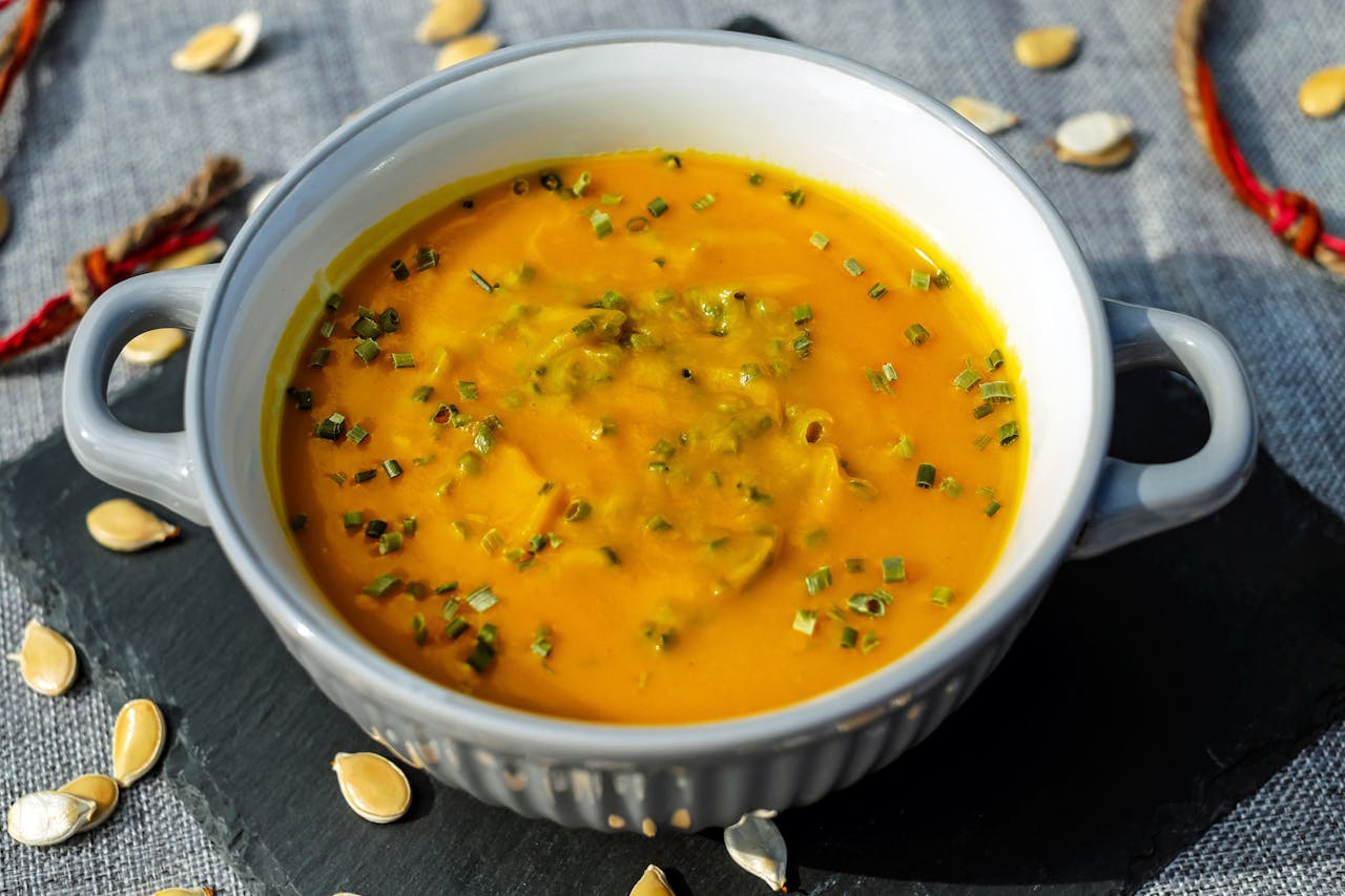 Bowl of creamy pumpkin or squash soup, topped with chopped chives, served in a grey dish with handles, surrounded by pumpkin seeds