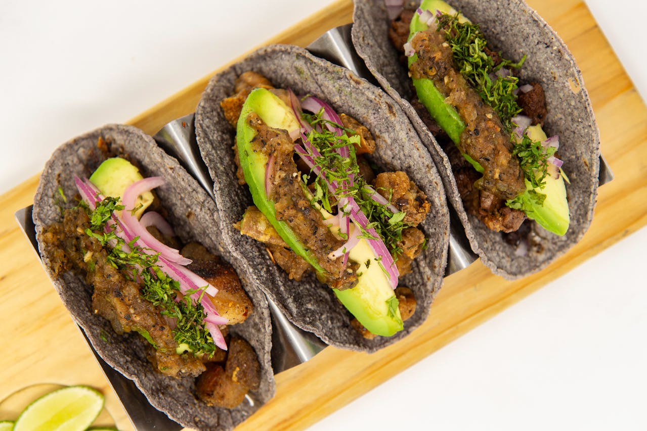 Three black bean tacos with blue corn tortillas, avocado slices, pickled onions, salsa, and fresh herbs on a wooden board