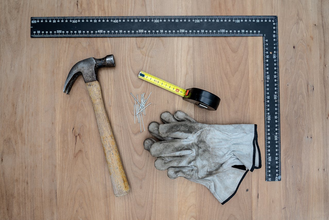 Claw hammer with wooden handle, tape measure extended, black electrical tape, large black carpenter’s square, all placed on a wooden surface