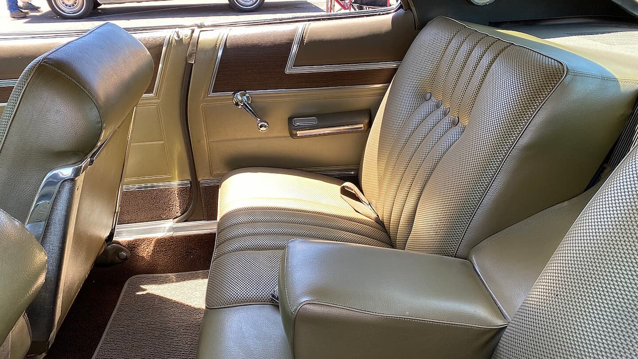 Green vinyl and fabric seats, sunlight casting strong shadows, chrome seat adjuster visible, manual window crank, brown carpet flooring, armrest between seats
