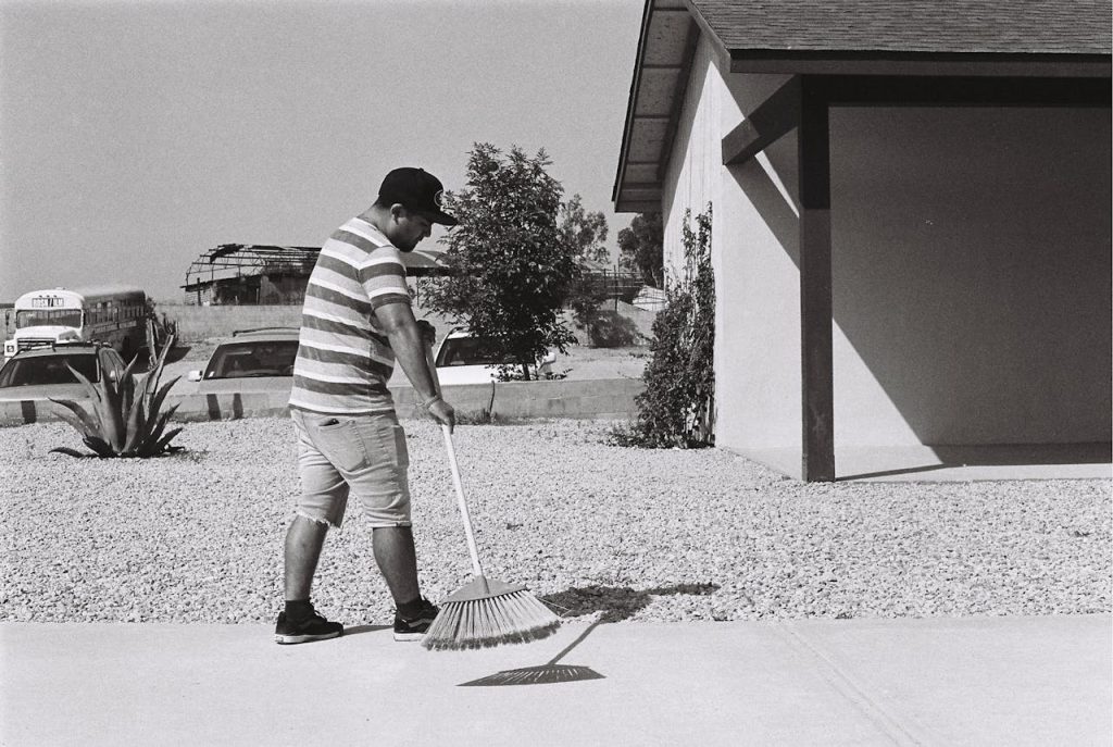 Man in striped shirt sweeping sidewalk, wearing cap and shorts, standing outside a building on a sunny day