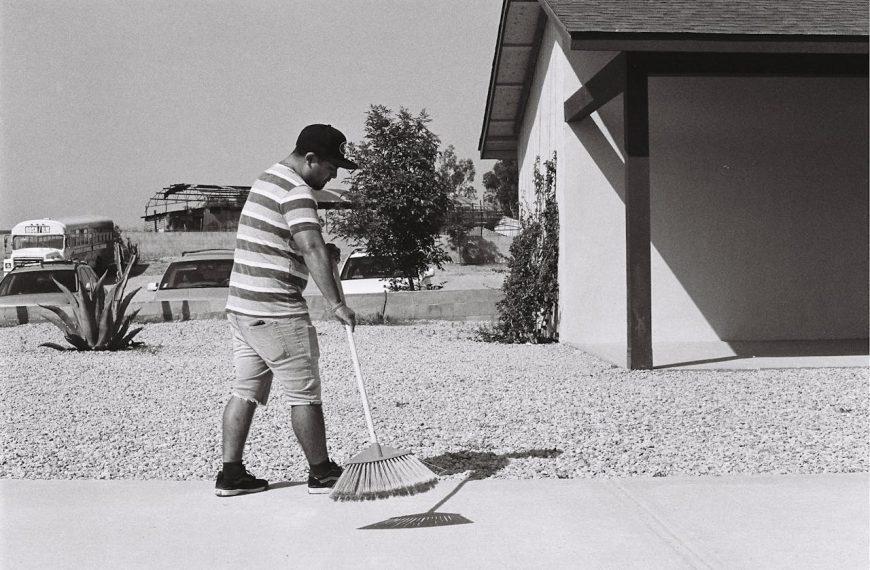 Man in striped shirt sweeping sidewalk, wearing cap and shorts, standing outside a building on a sunny day