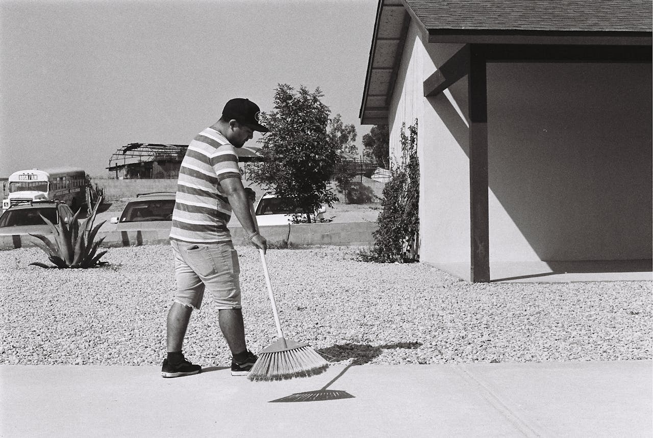 Man in striped shirt sweeping sidewalk, wearing cap and shorts, standing outside a building on a sunny day