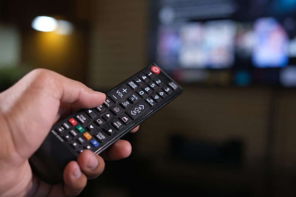 hand holding black TV remote control, pointing at blurred television screen in background, buttons clearly visible, indoor setting