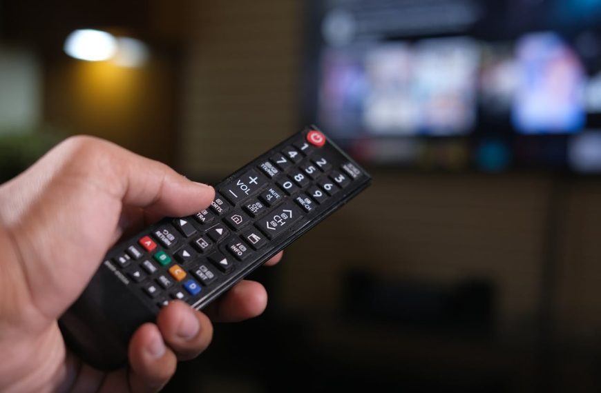 hand holding black TV remote control, pointing at blurred television screen in background, buttons clearly visible, indoor setting
