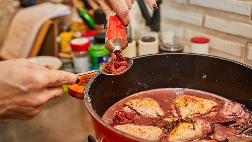 Chef adds tomato paste to rosemary-marinated chicken in pot on gas stove.