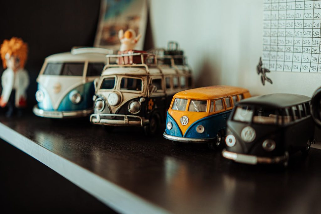 Row of vintage Volkswagen van toys, various colors including blue, yellow, black, and white, placed on a dark wooden shelf, background includes a grid chart and small figurines