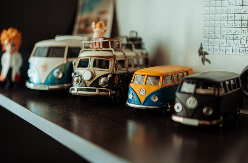 Row of vintage Volkswagen van toys, various colors including blue, yellow, black, and white, placed on a dark wooden shelf, background includes a grid chart and small figurines