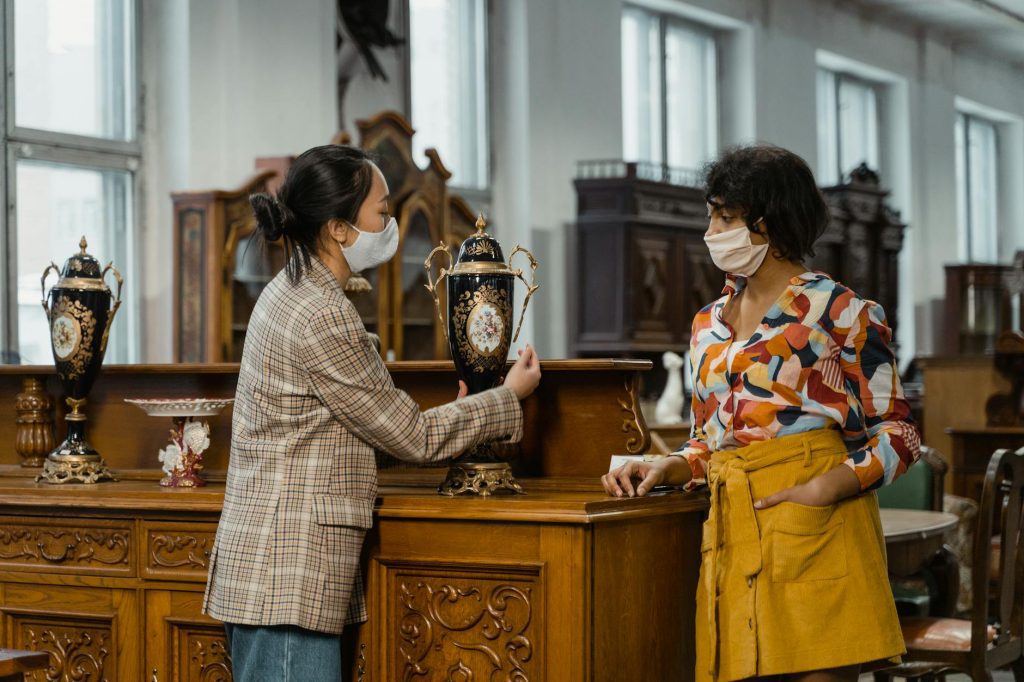 Two women wearing face masks examining an antique vase in a vintage furniture store, surrounded by wooden cabinets and ornate decor