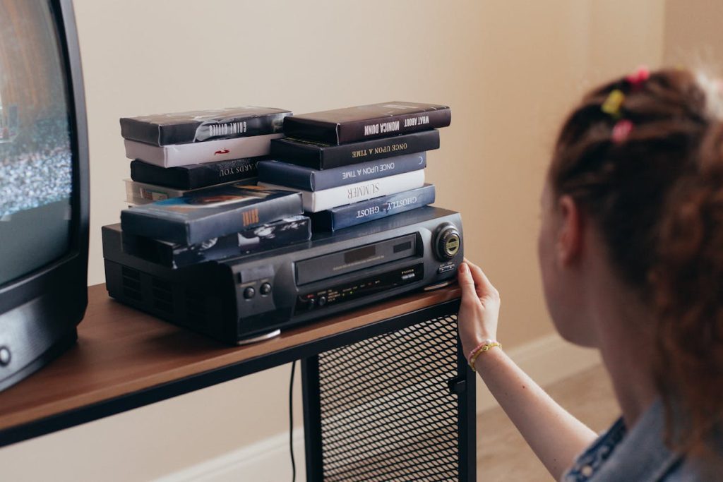 Girl operating VHS player, stack of VHS tapes on top, old TV with static screen, wooden and metal stand, cozy indoor setting