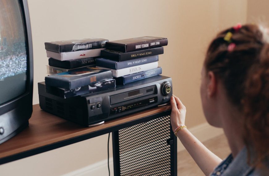 Girl operating VHS player, stack of VHS tapes on top, old TV with static screen, wooden and metal stand, cozy indoor setting